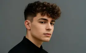 Młody mężczyzna z kręconymi, puszystymi włosami (fryzura fluffy hair) i wygolonymi bokami, ubrany w czarną koszulę, na szarym tle.